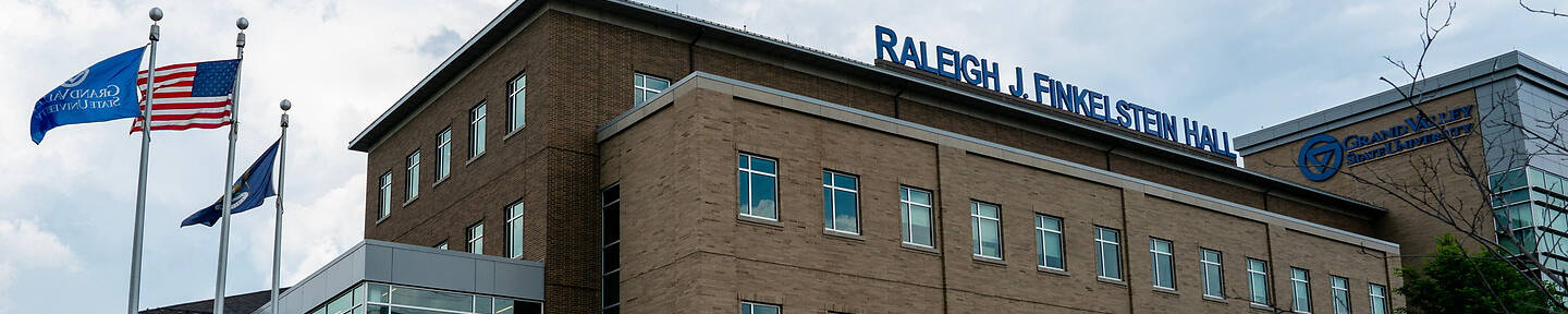 Raleigh J. Finkelstein Hall at Grand Valley State University is a modern brick building with flags flying outside and a paved road in front.